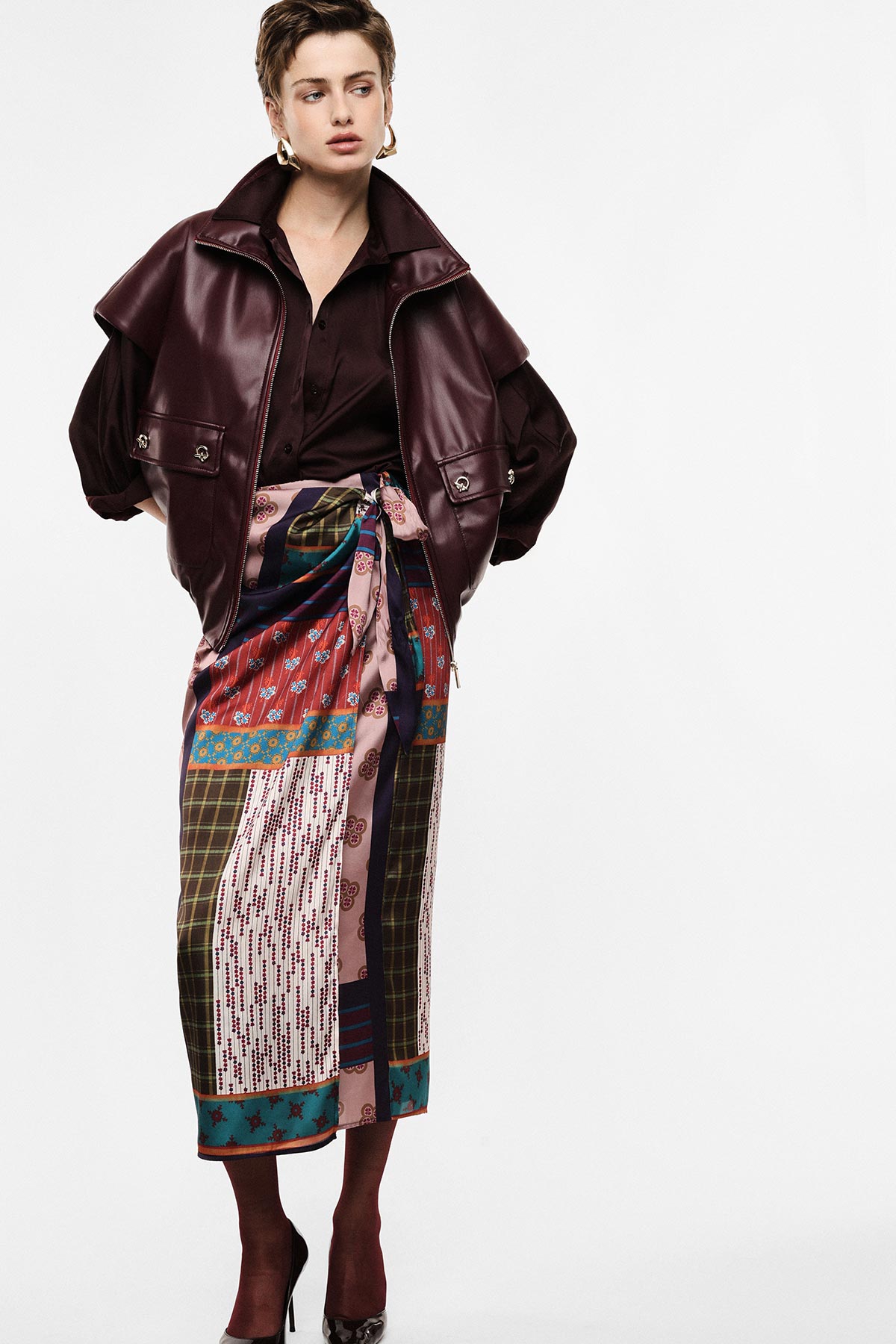 Woman modelling burgundy leather jacket, patterned patchwork wrap skirt and burgundy heels against plain white background
