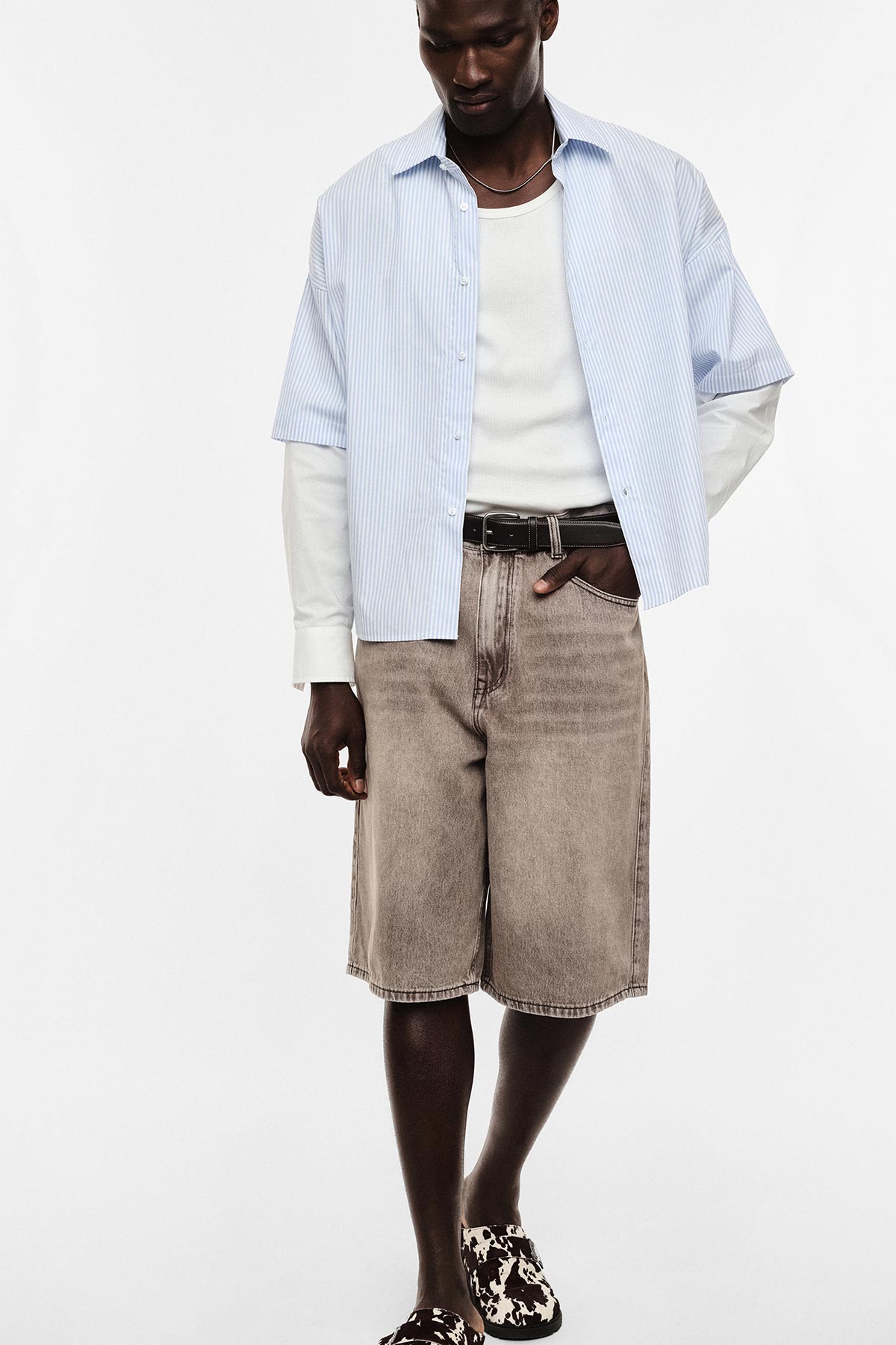 Man wearing blue striped short-sleeve shirt over white top, grey denim knee-length shorts and black-white patterned slip-ons