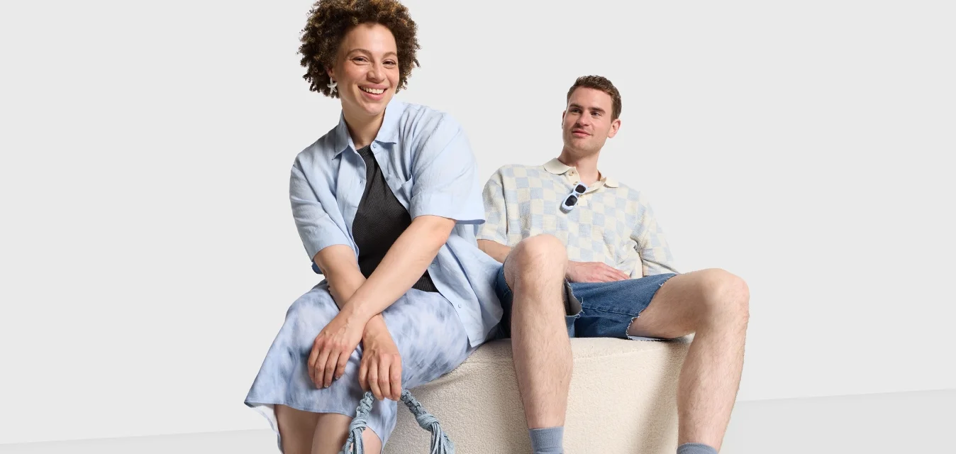 Due persone siedono su un pouf rotondo beige con una texture. Una indossa una camicia azzurro chiaro, una gonna di jeans e delle scarpe flat blu con borchie; l'altra indossa una camicia con motivo e dei pantaloni shorts in denim.