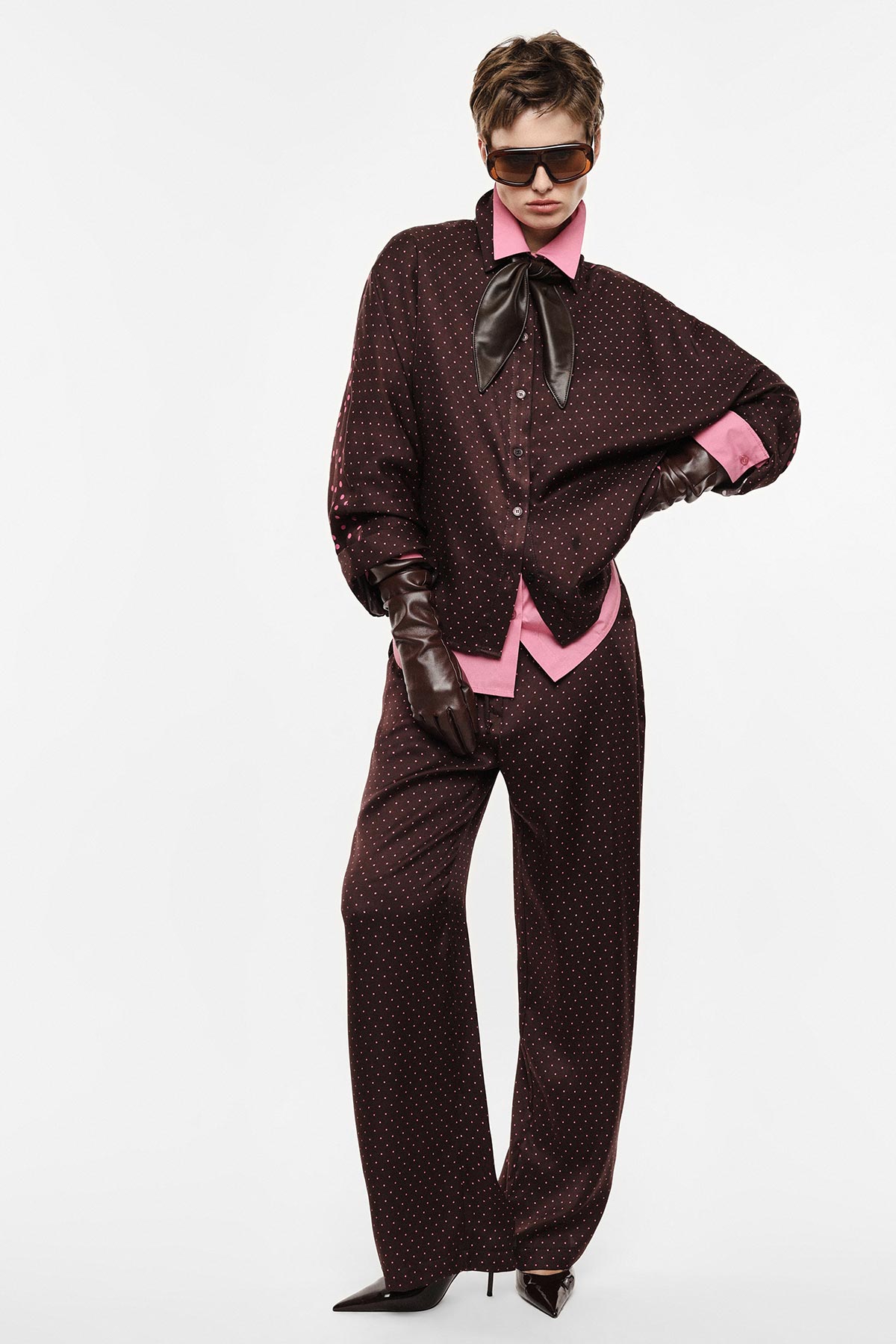 Fashion model in dark brown polka-dot oversized suit with pink shirt, leather bow and gloves, sunglasses and heels on white background