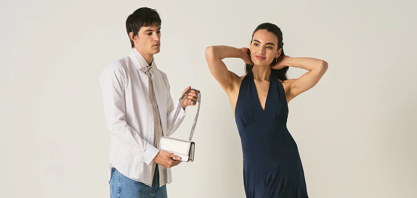 Uomo con camicia bianca e jeans che tiene una piccola borsa a mano accanto a una donna in abito blu navy che posa con le mani dietro la testa.
