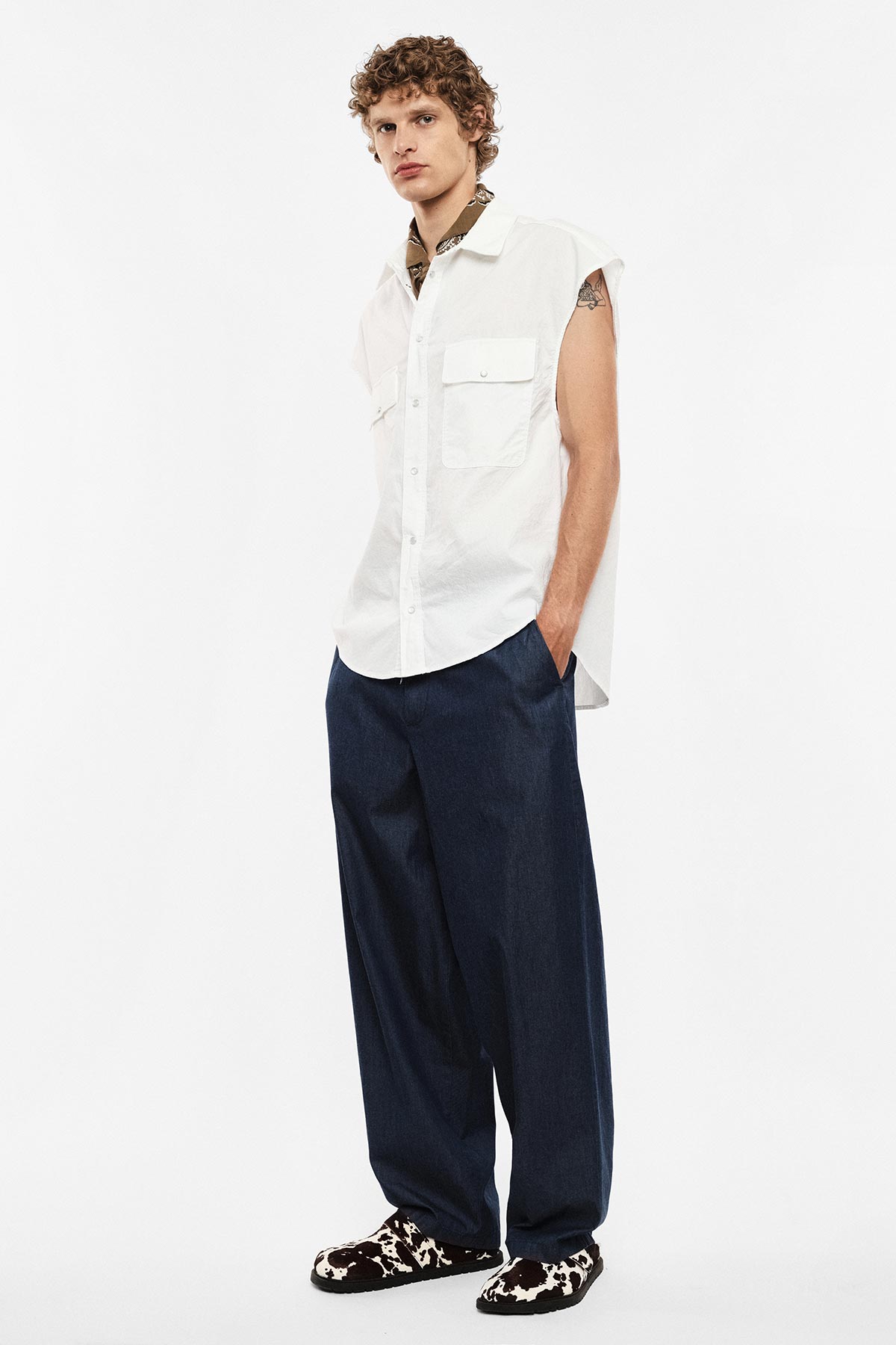 Man wearing sleeveless white button shirt, wide-leg navy trousers and patterned loafers, hands in pockets
