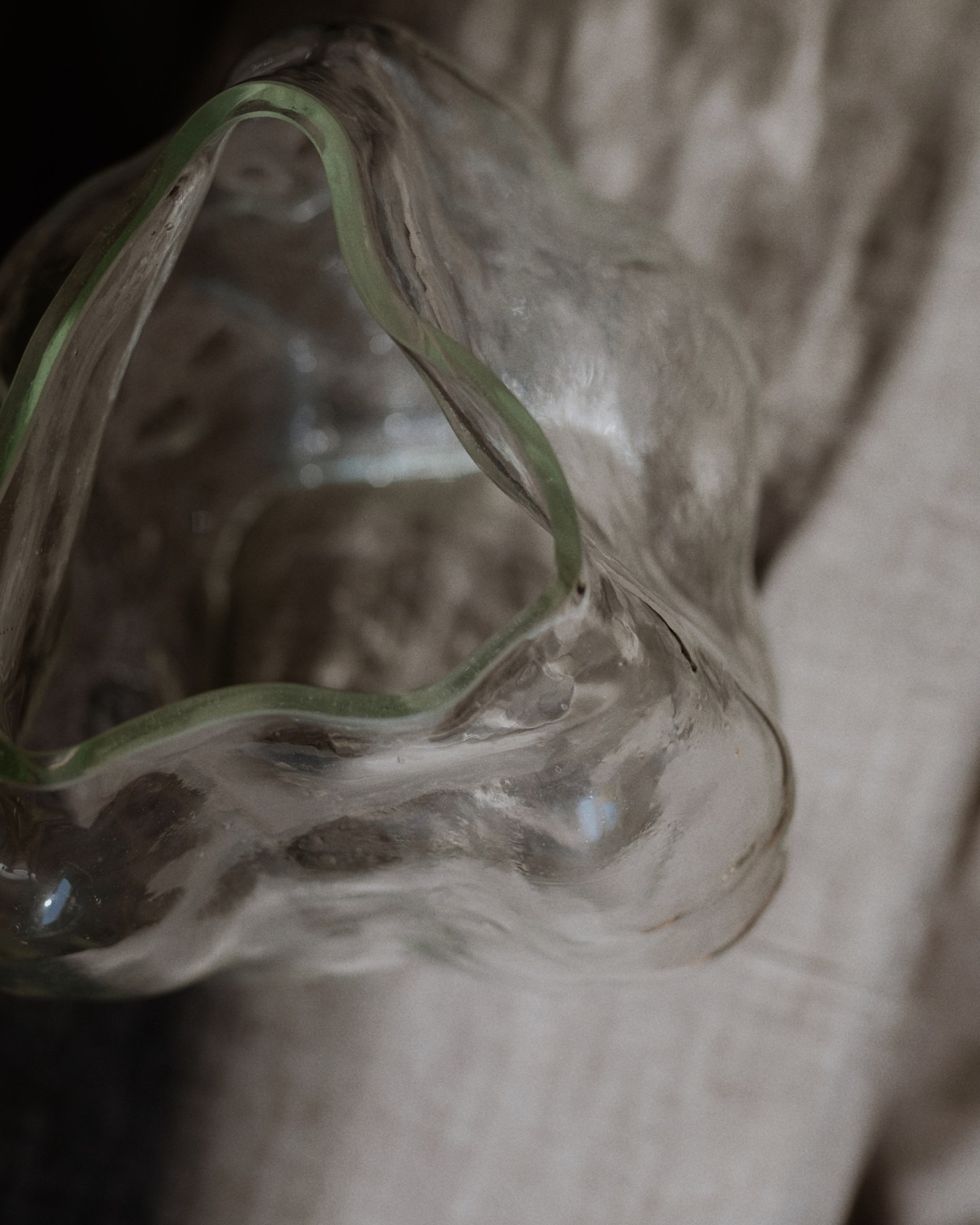 Close-up of a wavy clear glass bowl with a pale green rim resting on textured fabric