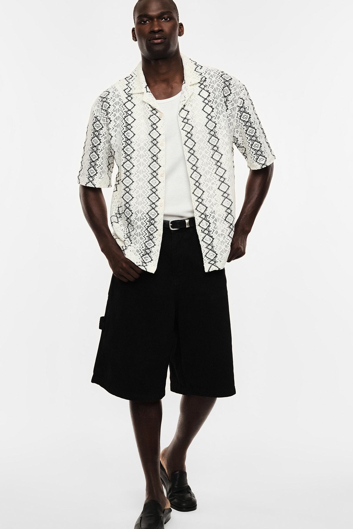 Man wearing white patterned short-sleeve shirt over white tee, black knee-length shorts and black loafers on white background