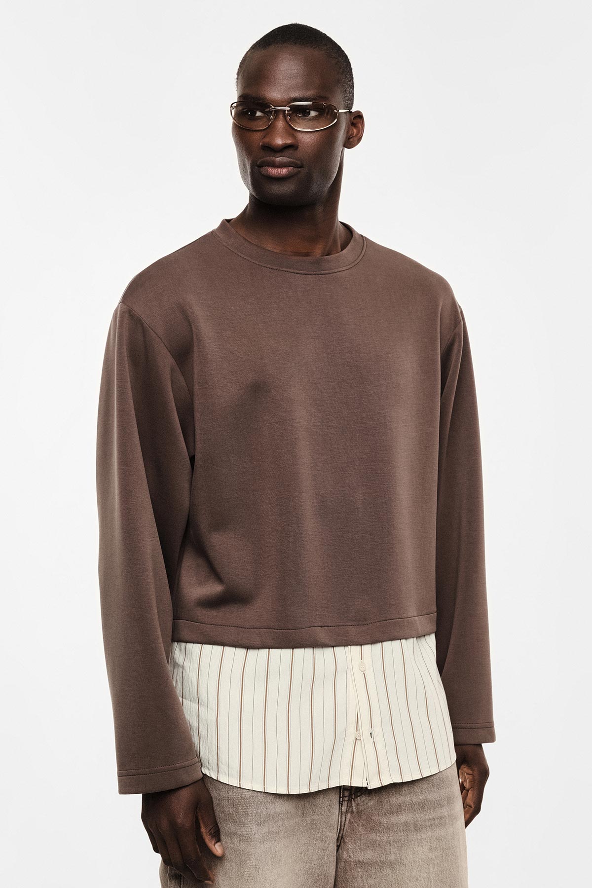 Male model wearing brown cropped sweatshirt over striped shirt, beige jeans and glasses on white background