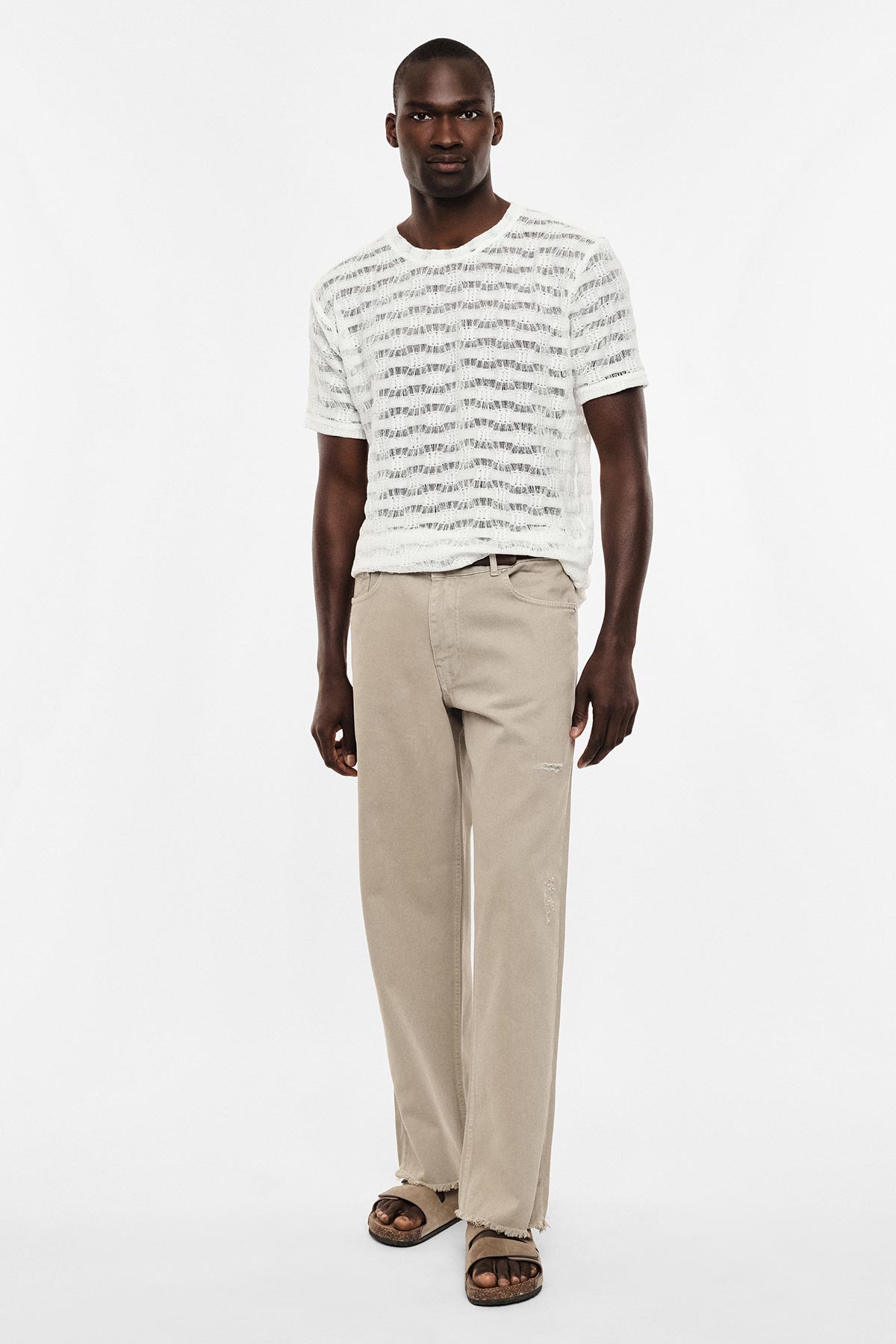 Male model wearing white textured short‑sleeve T-shirt, beige relaxed straight‑leg trousers and brown sandals, standing against a white background