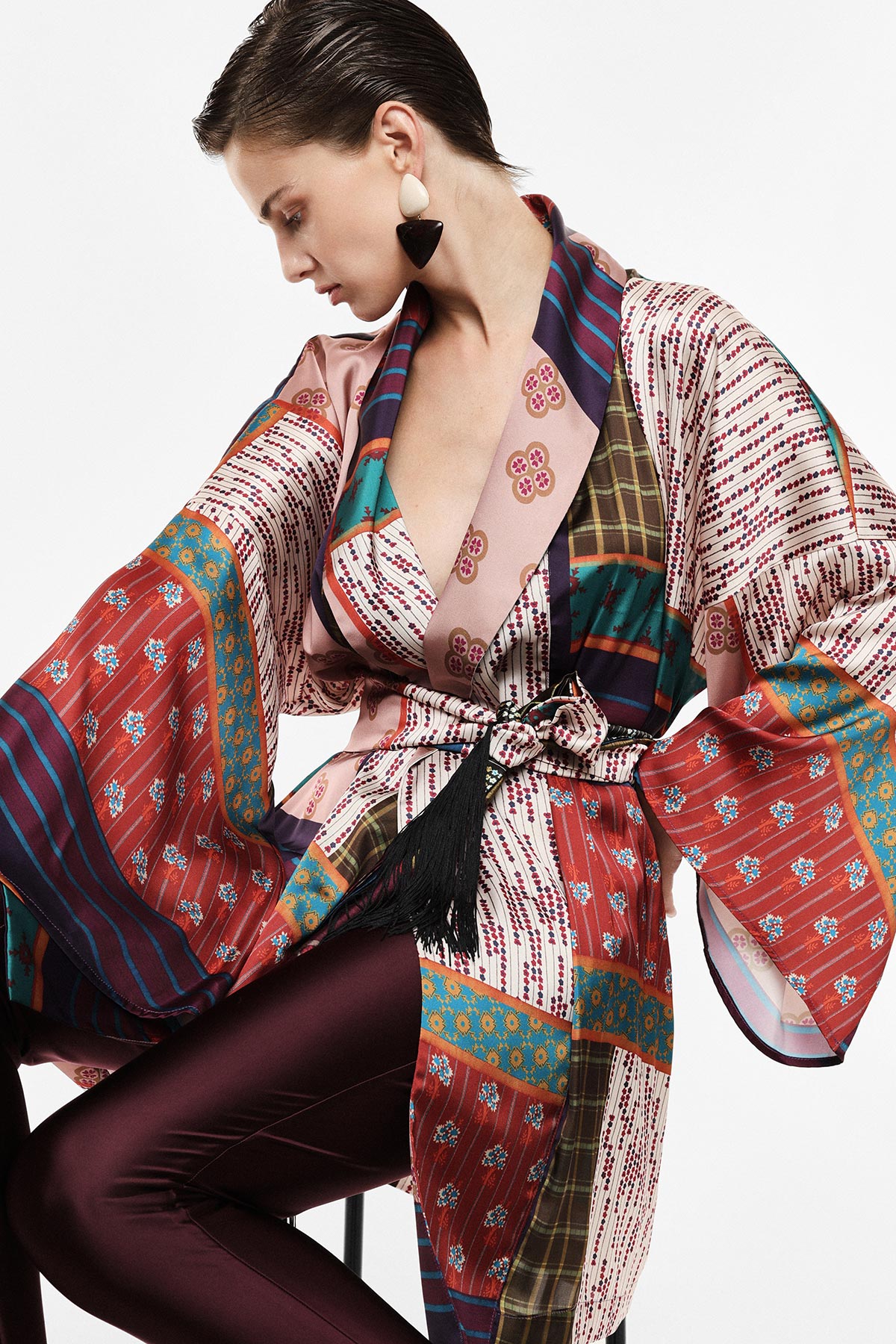 Woman wearing a colourful patterned silk kimono robe with wide sleeves, tasseled belt and burgundy satin trousers, seated in profile