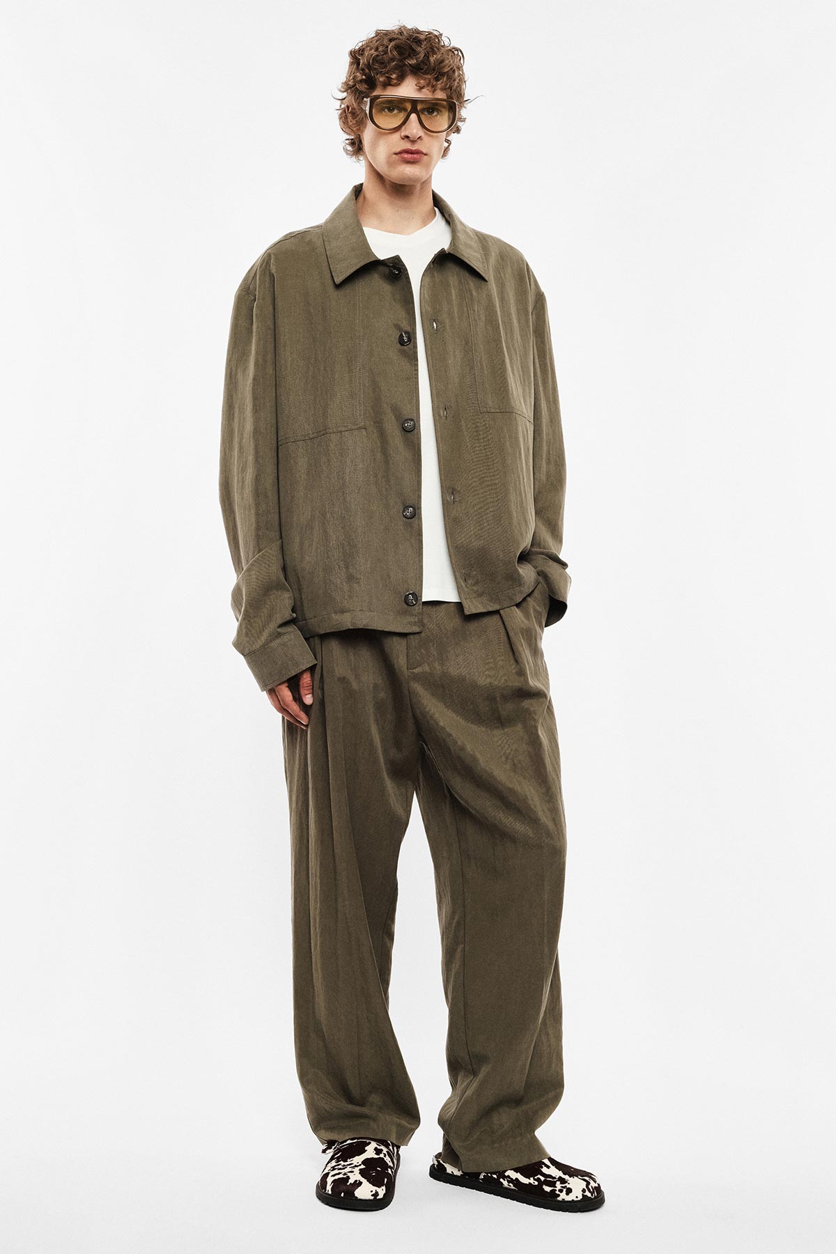 Person wearing olive corduroy relaxed-fit jacket and wide trousers, white T-shirt, retro tinted glasses and cow-print loafers.