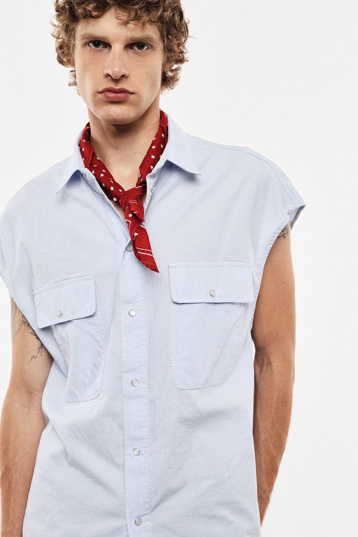 Young man wearing sleeveless pale blue shirt and red bandana neck scarf, cropped portrait with visible arm tattoos