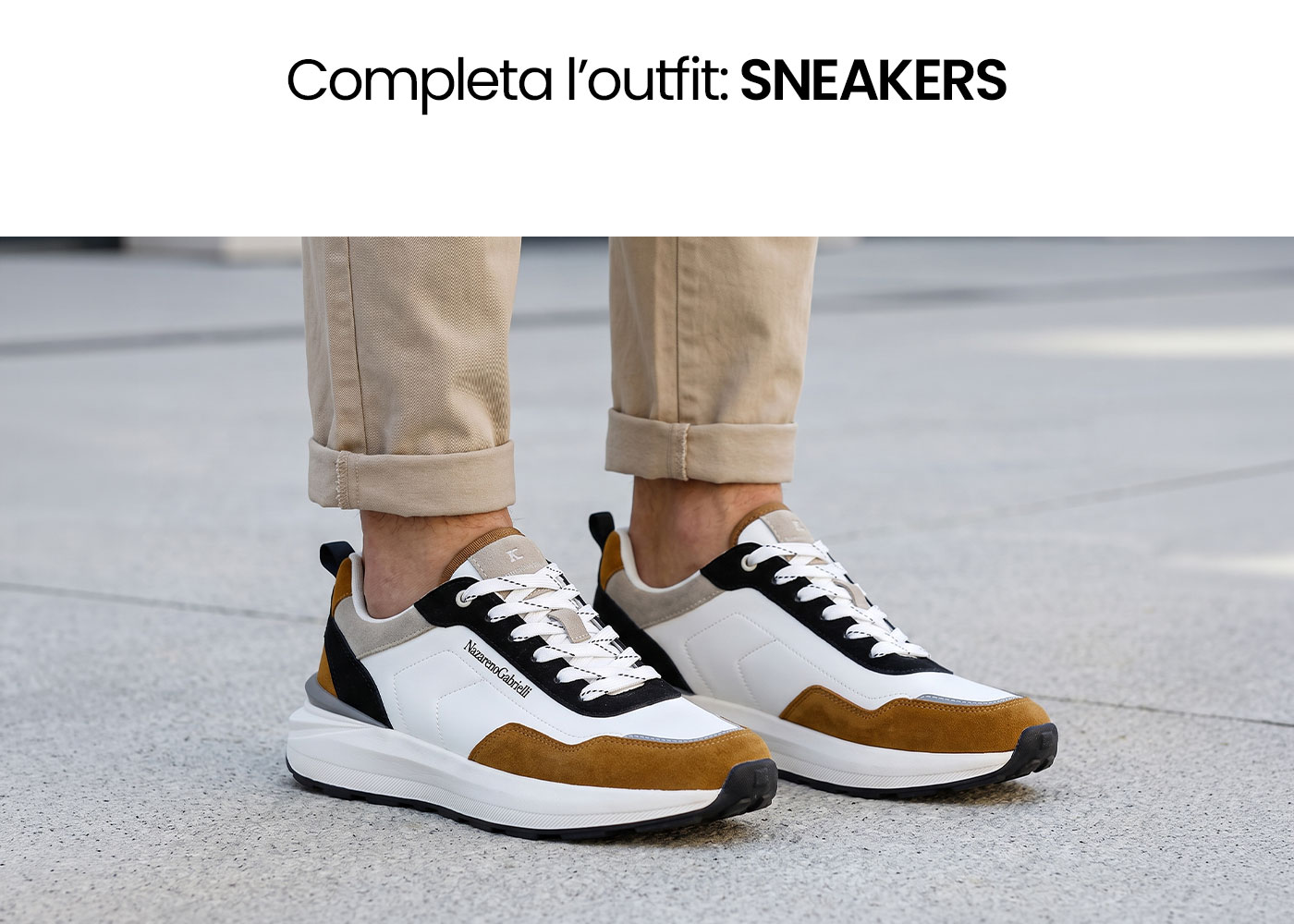 Pair of white, black and tan suede sneakers on feet with rolled-up beige chinos standing on urban pavement