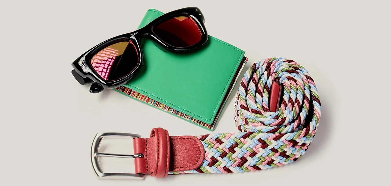 Occhiali da sole neri su un portafoglio verde accanto a una cintura intrecciata multicolore arrotolata con fibbia in pelle rossa su sfondo chiaro.