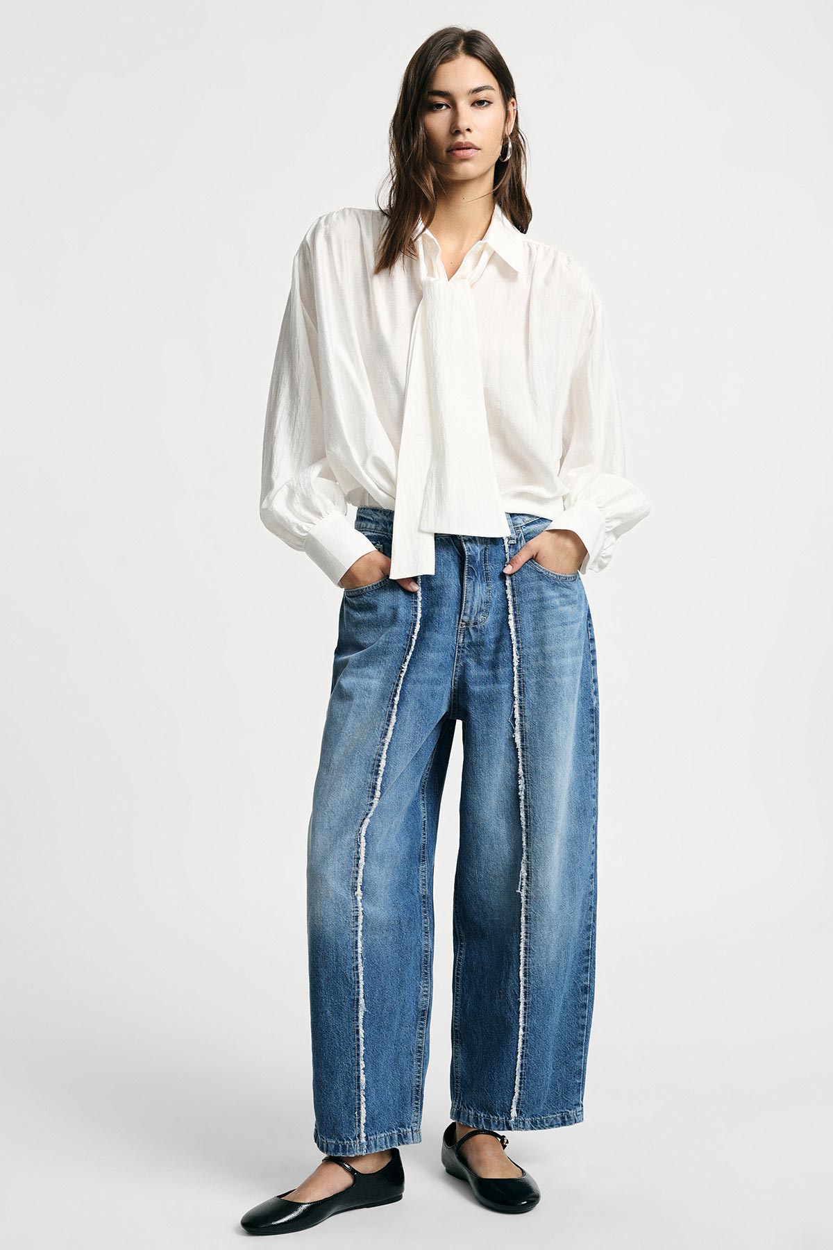 Woman modelling white tie-neck blouse and wide-leg blue jeans with exposed vertical seams, black flats, hands in pockets