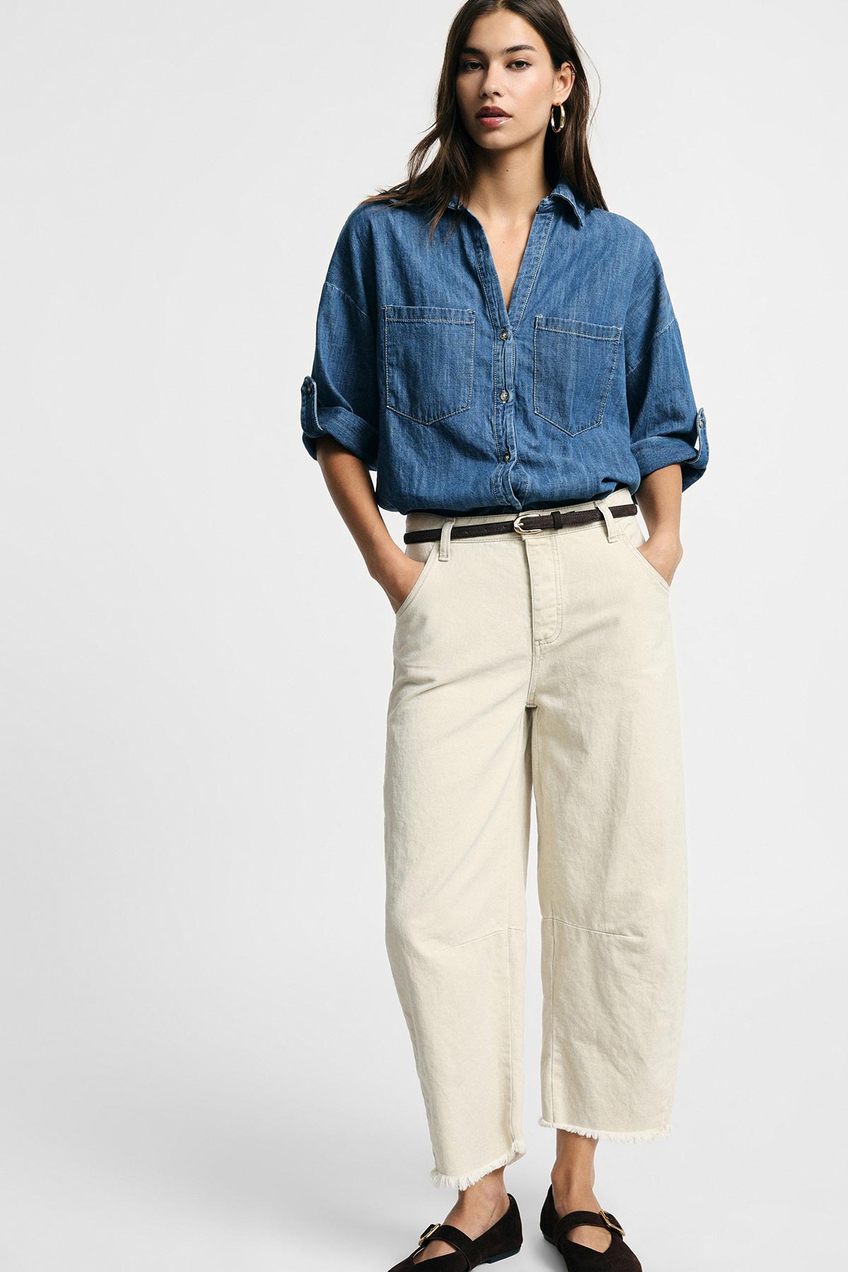 Woman modelling a blue denim shirt with cream wide-leg cropped trousers, brown belt and black loafers in a studio portrait