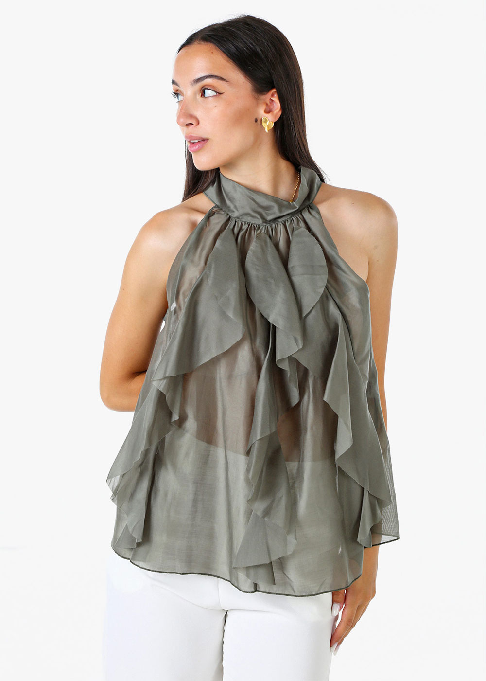 Woman wearing sheer olive-green halter ruffle blouse and white trousers, standing and looking left