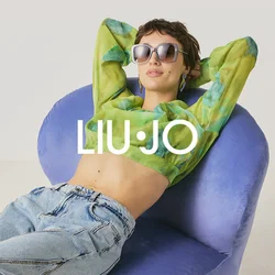 Persona con occhiali da sole, top corto verde a motivo e jeans che si rilassa su una sedia moderna viola contro uno sfondo chiaro e uniforme.