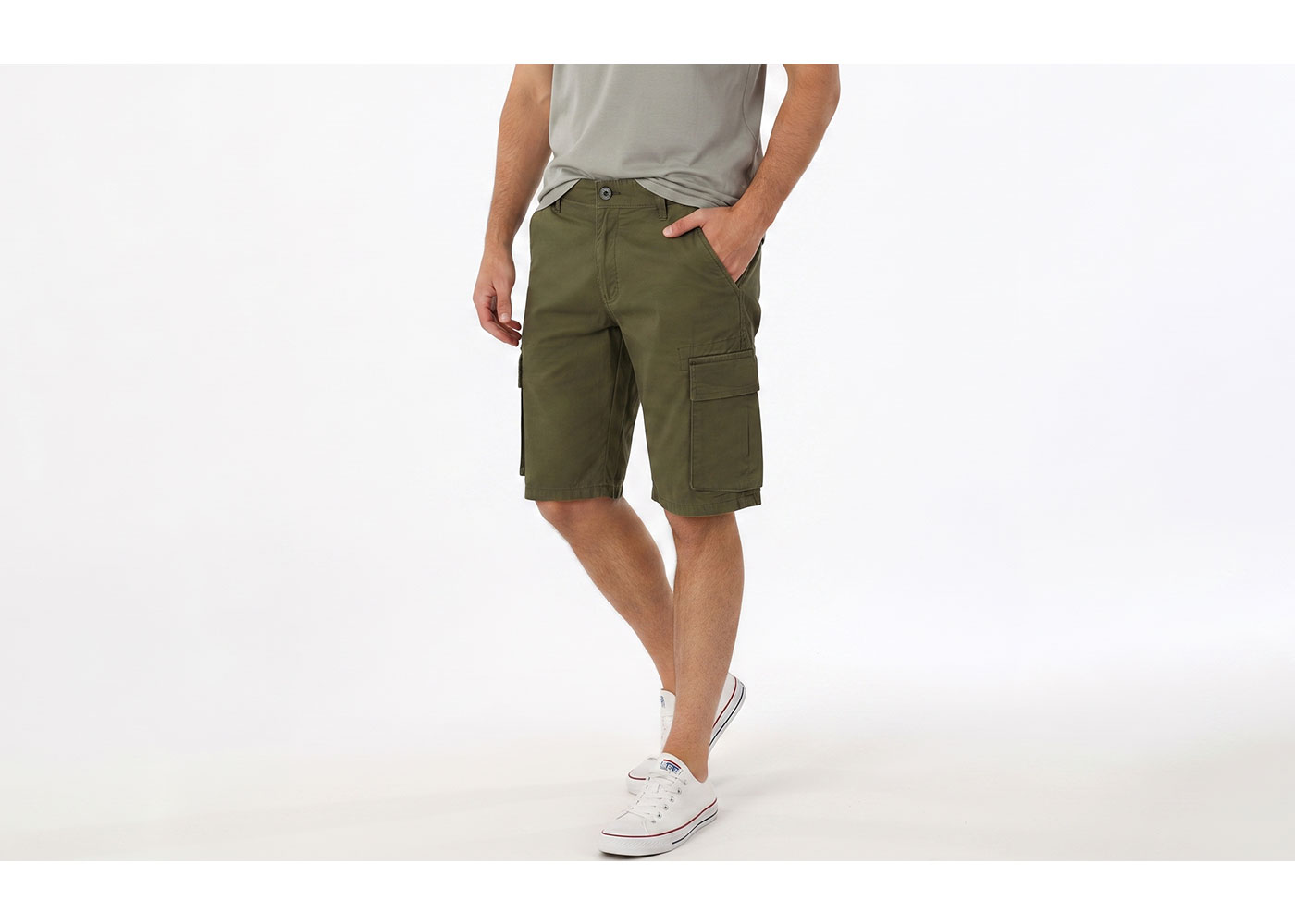 Male model wearing olive green cargo shorts with side pockets and white canvas trainers, casual standing pose on plain white background