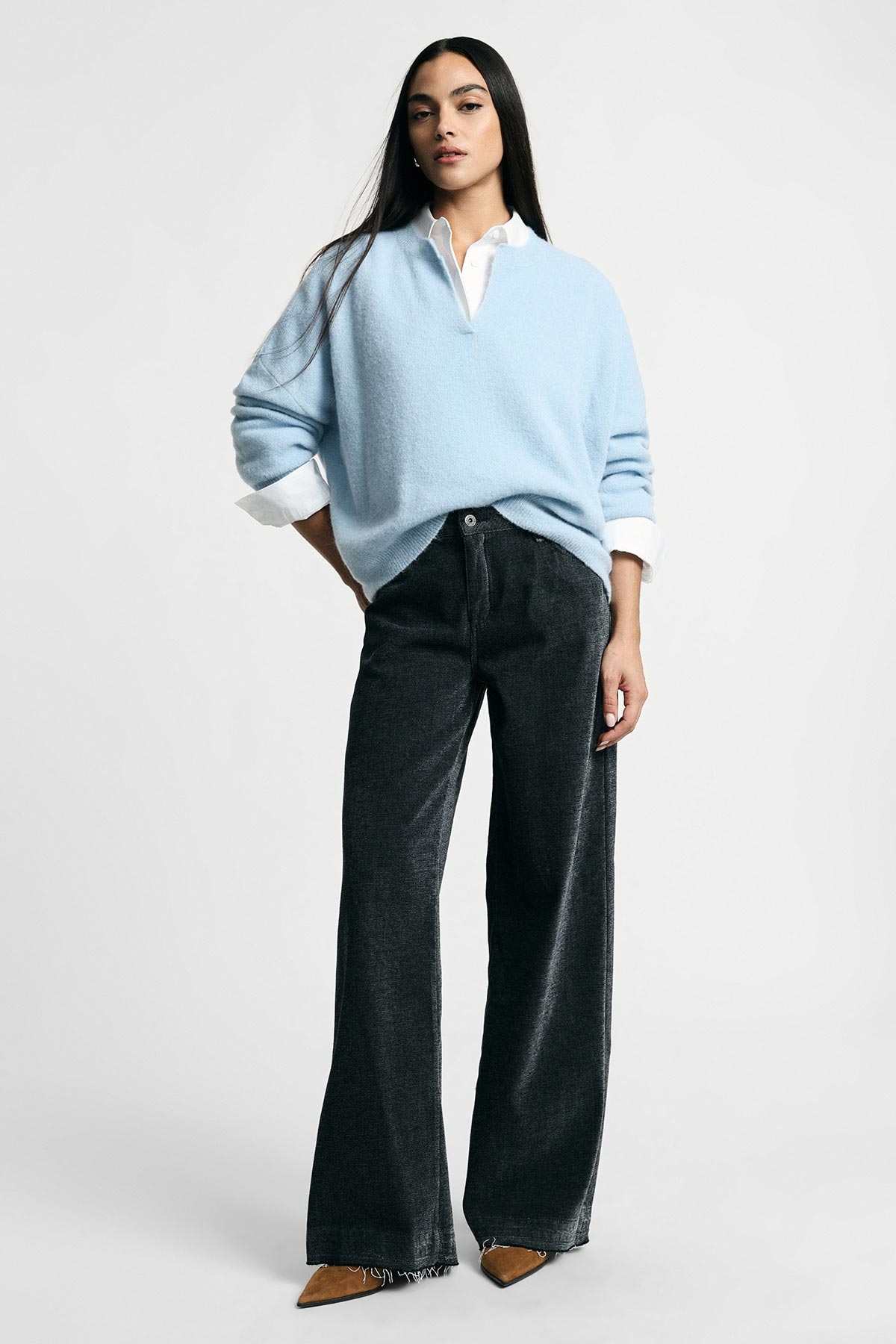 Woman standing against plain background wearing a light blue V-neck sweater over a white shirt, black wide-leg trousers and brown pointed shoes