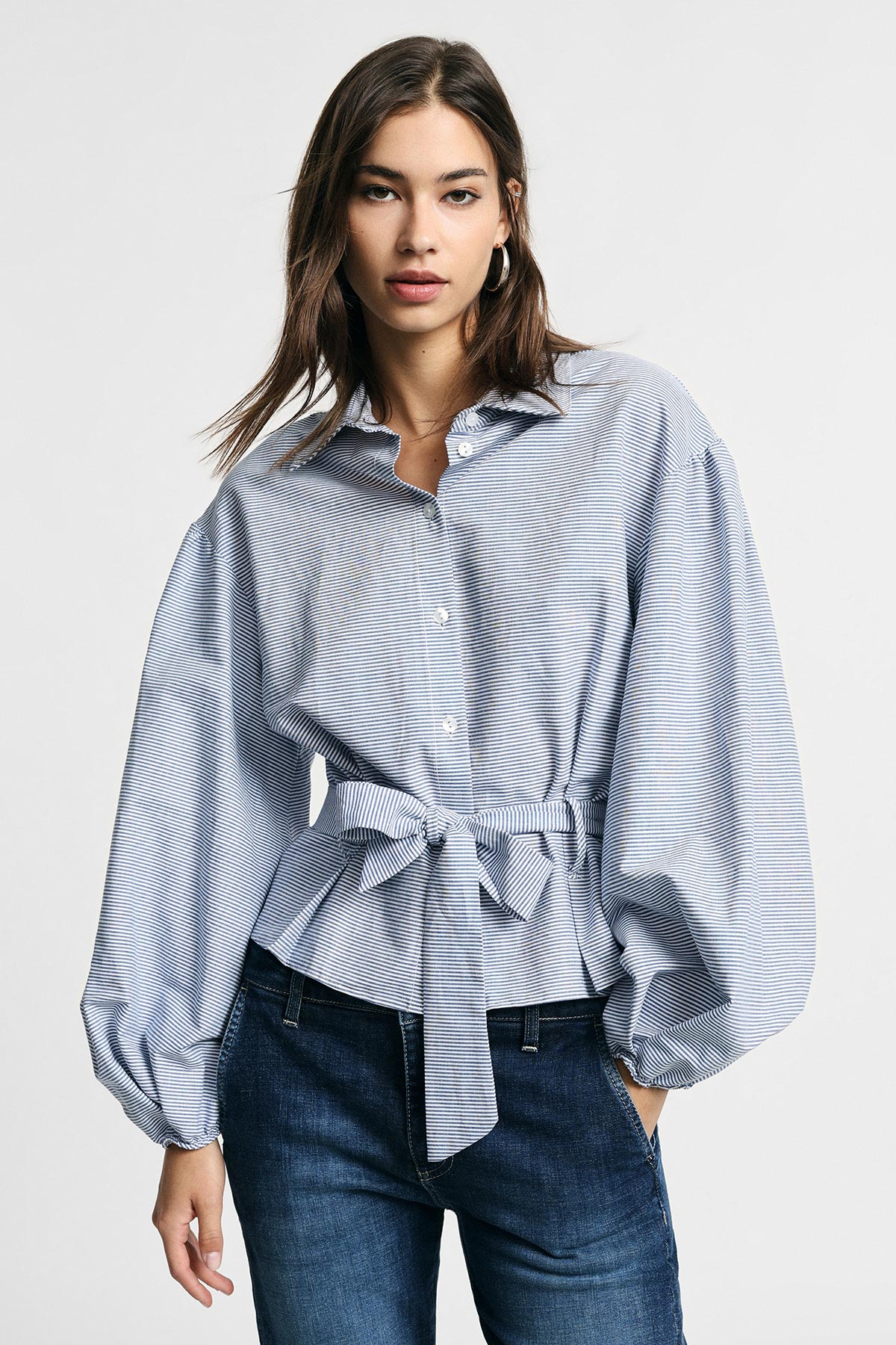 Woman in blue striped tie‑waist blouse with balloon sleeves paired with dark jeans against white background