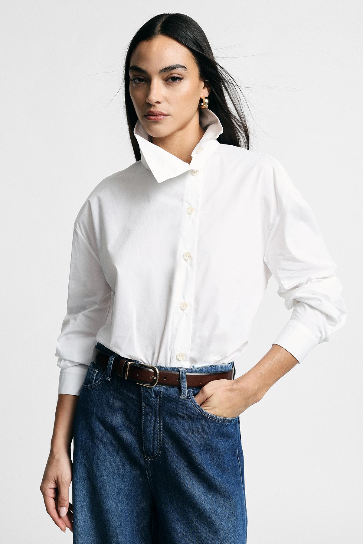 Woman modelling white button-up shirt tucked into denim jeans with brown belt, gold hoop earring and hand in pocket on plain background