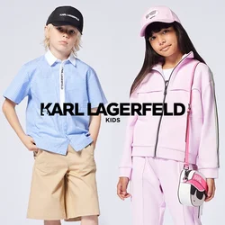 Due bambini stanno in piedi; uno indossa una camicia a righe blu con pantaloncini beige e un berretto nero, l'altro una tuta rosa e un cappello con una piccola borsa.