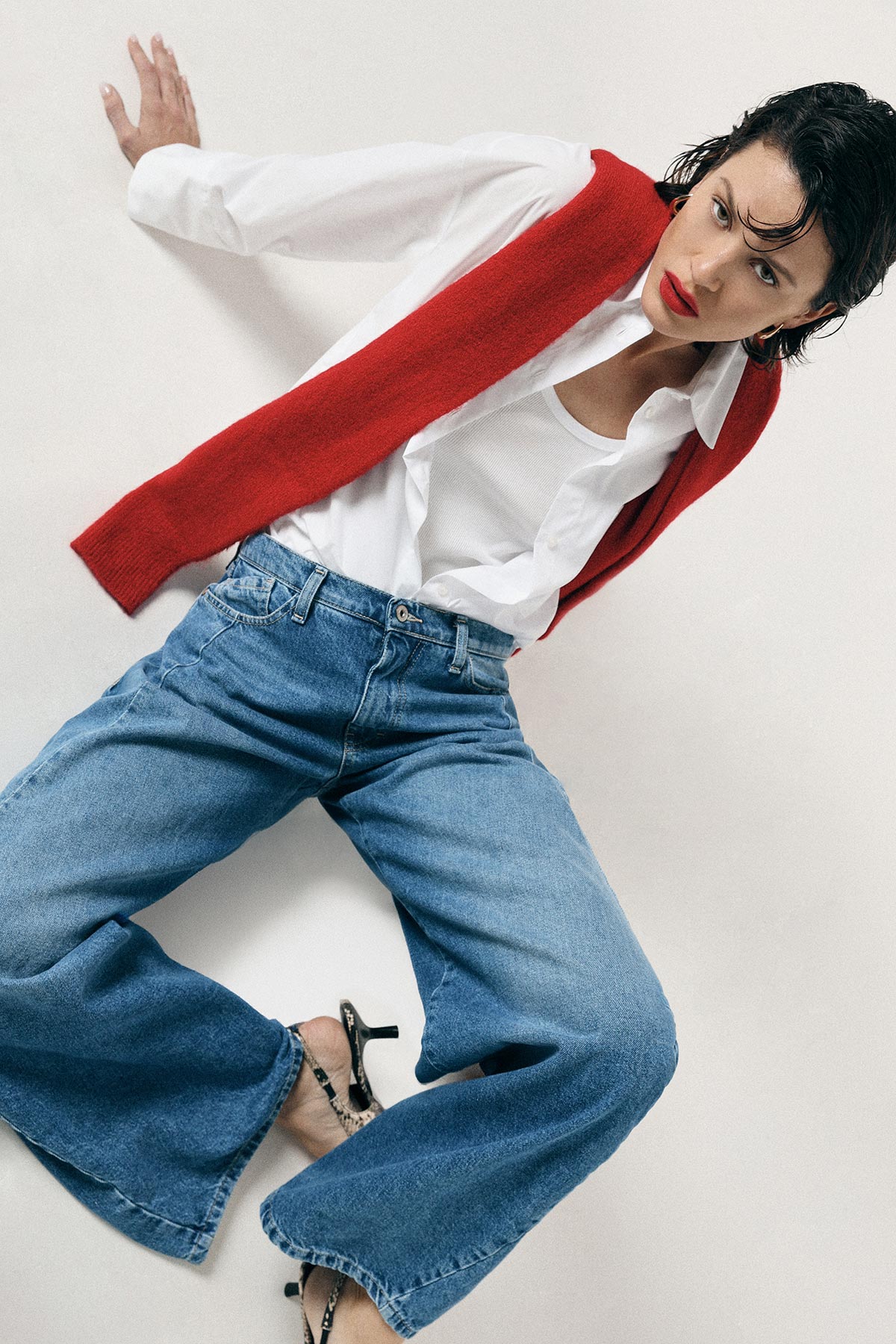 Woman modelling red sweater draped over shoulders, white shirt and wide-leg jeans with heels in studio pose