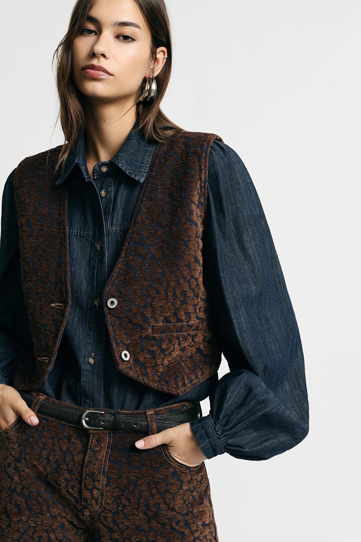 Woman wearing dark denim shirt with brown patterned vest and matching trousers, leather belt, hoop earring