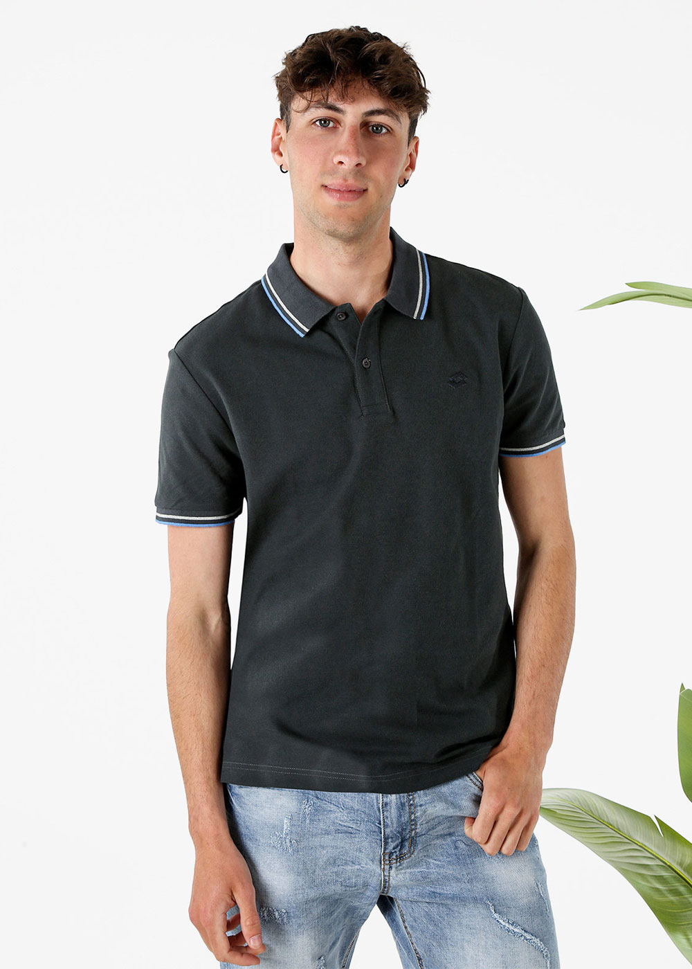Young man wearing a black short-sleeve polo with blue-trim collar and faded blue jeans, standing with hand in pocket against a white background