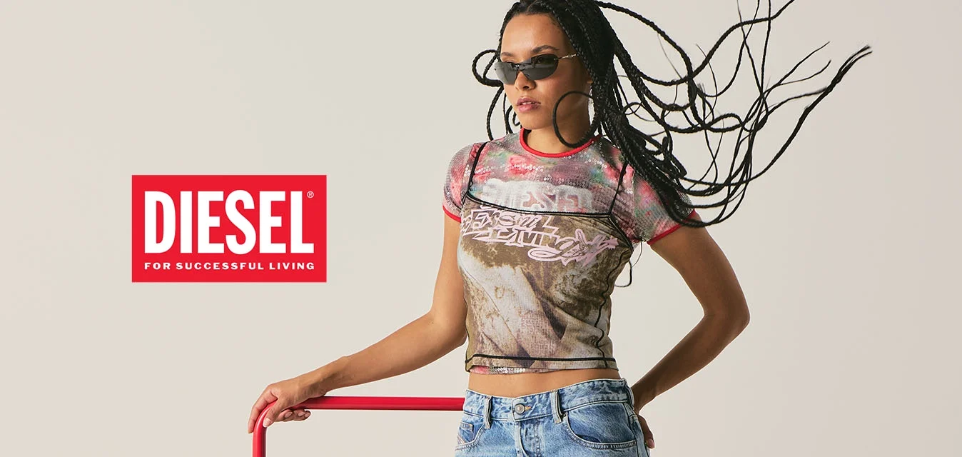 Donna con capelli intrecciati e occhiali da sole, che indossa una maglietta grafica e jeans, appoggiata a una struttura metallica rossa contro uno sfondo semplice.