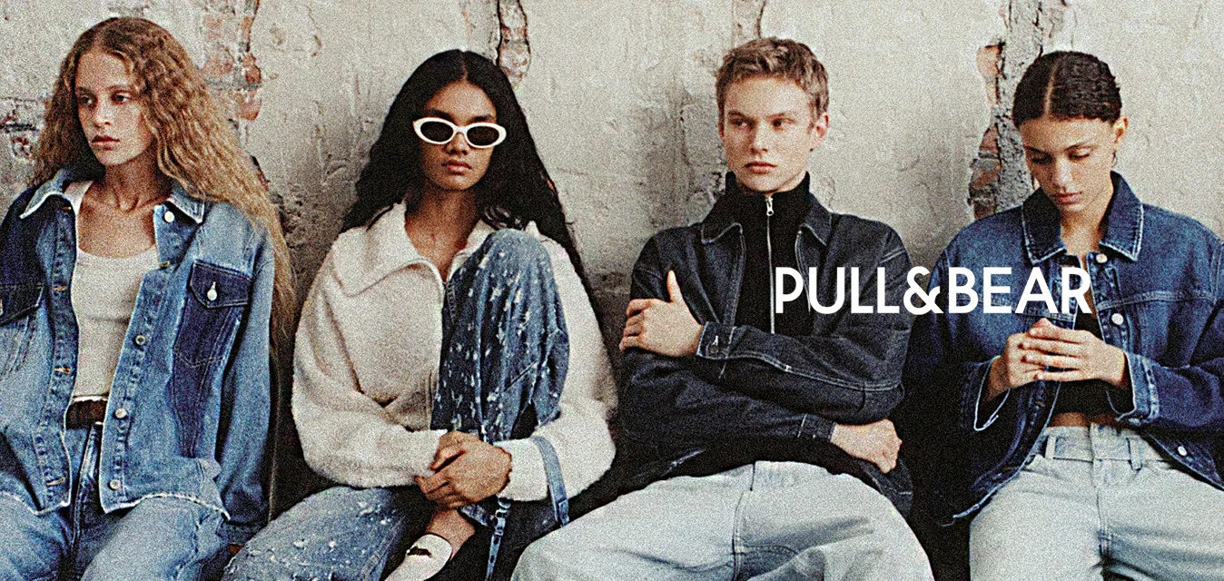 Sei giovani adulti che indossano abbigliamento di denim oversize siedono contro un muro di mattoni usurato, alcuni usano il telefono e altri guardano avanti.