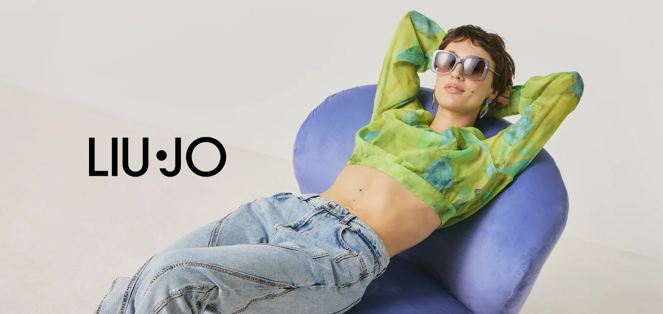 Persona con occhiali da sole, top corto verde a motivo e jeans che si rilassa su una sedia moderna viola contro uno sfondo chiaro e uniforme.