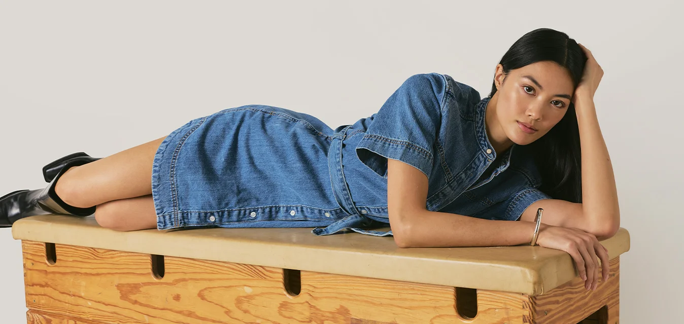 Donna con un vestito di jeans blu sdraiata di lato su una cassa di legno da palestra, con la testa appoggiata su una mano, guarda verso la fotocamera.