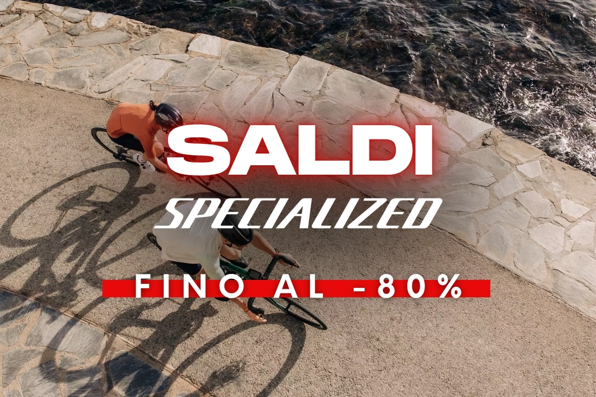 Aerial view of two cyclists riding along a stone seaside promenade, with red text overlay advertising 'SALDI Specialized fino al -80%'