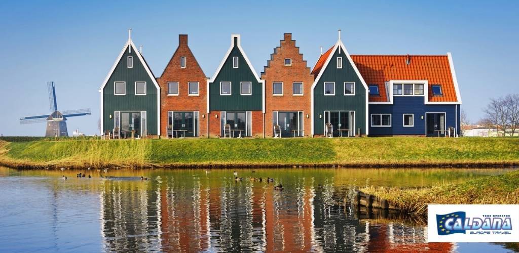 Colourful Dutch canal houses with stepped gables and red-tiled roofs beside a windmill, reflected in calm water with blue sky