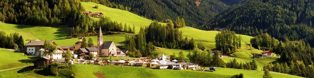 Alpine village in a sunlit green valley with a stone church steeple, timber houses and forested hills leading to mountains