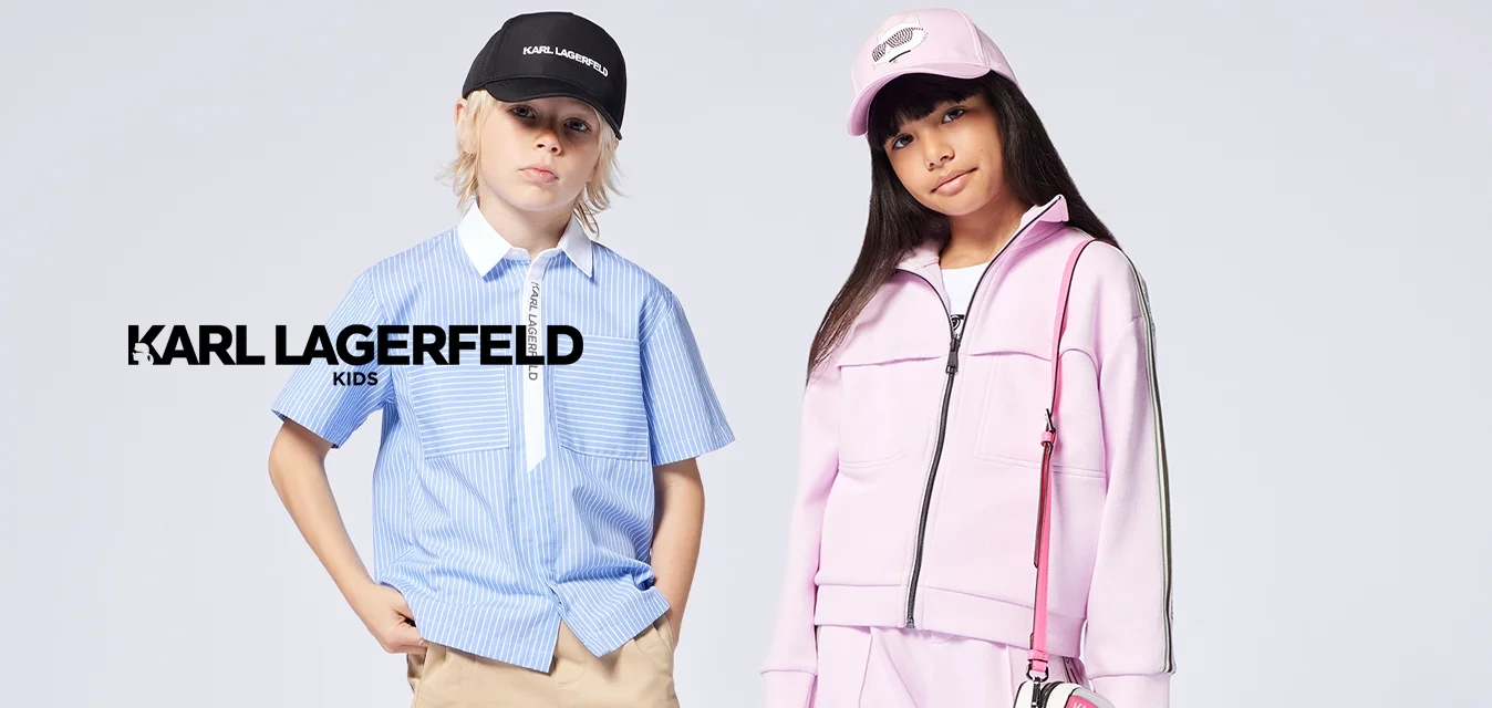 Due bambini stanno in piedi; uno indossa una camicia a righe blu con pantaloncini beige e un berretto nero, l'altro una tuta rosa e un cappello con una piccola borsa.