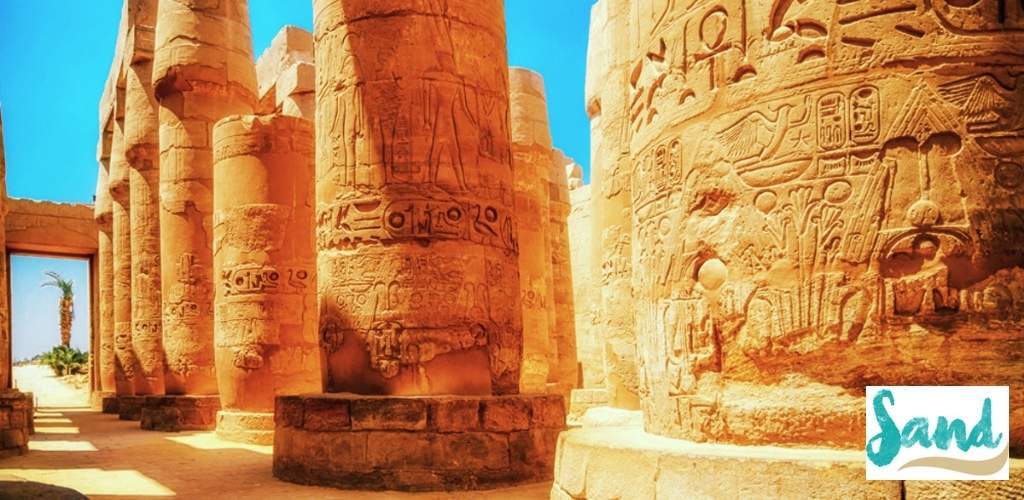 Sunlit row of ancient Egyptian temple columns carved with hieroglyphs and reliefs, sandy courtyard and palm visible through doorway