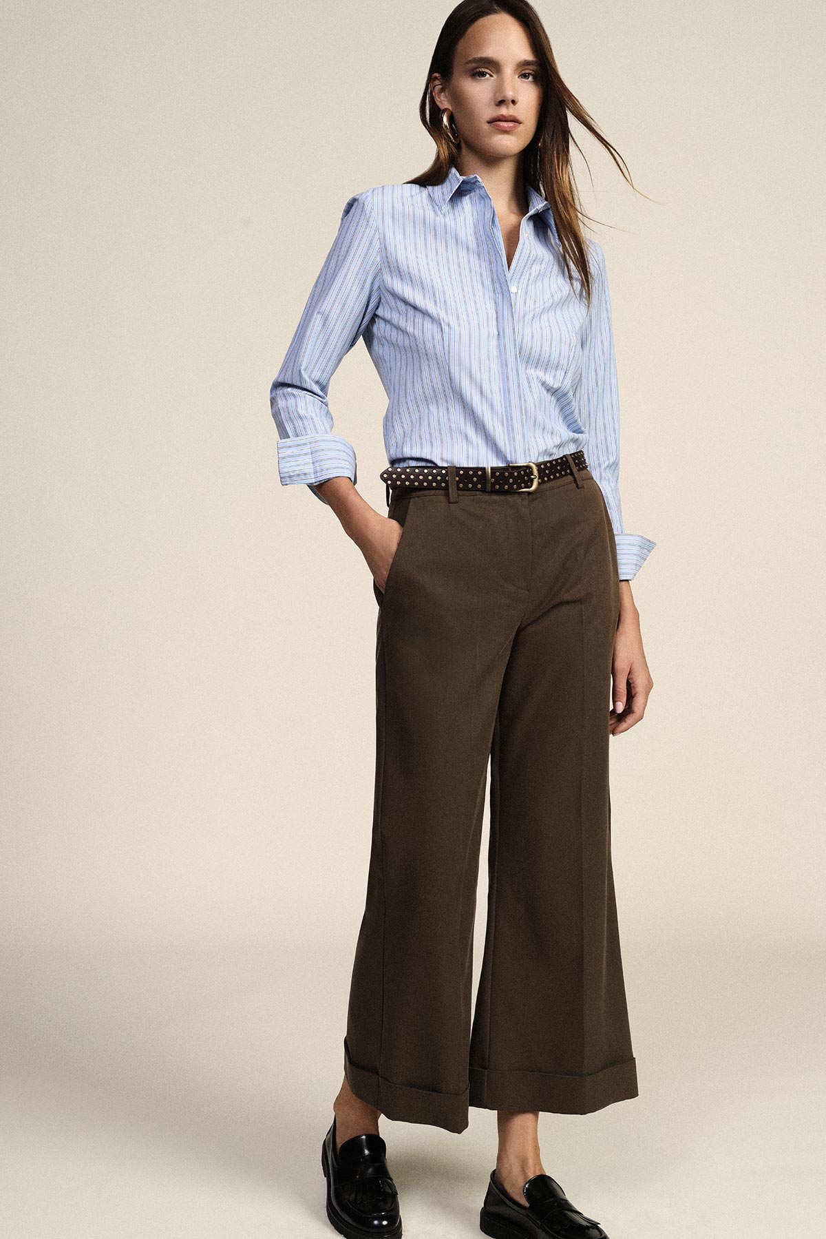 Woman modelling blue striped shirt tucked into high-waist brown wide-leg trousers with studded belt and loafers