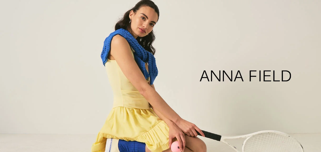 Donna in vestito giallo inginocchiata, che tiene una pallina da tennis rosa e una racchetta da tennis bianca su uno sfondo chiaro e uniforme.