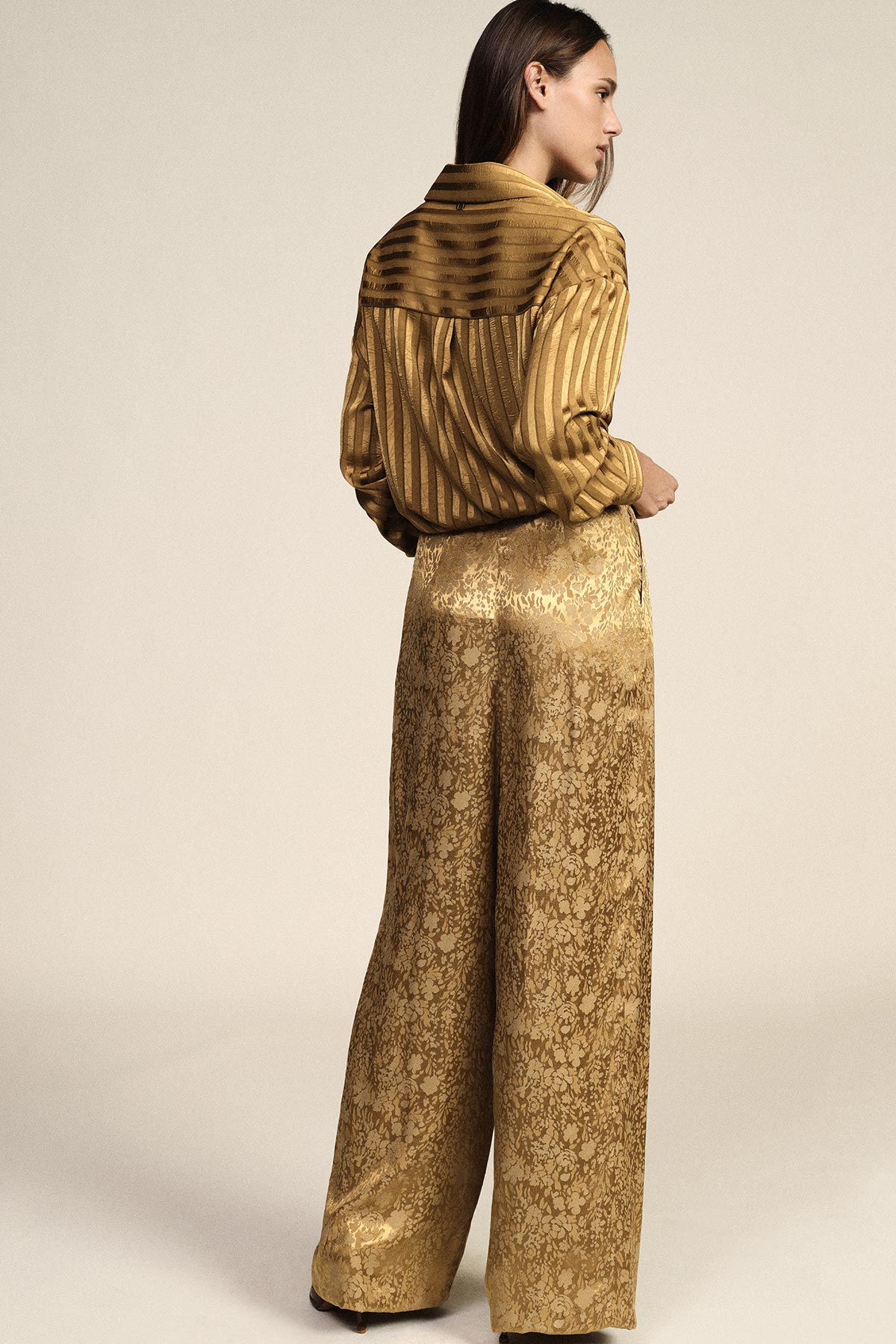 Woman seen from behind wearing a gold striped silk blouse and high-waisted gold brocade wide-leg trousers against a neutral background