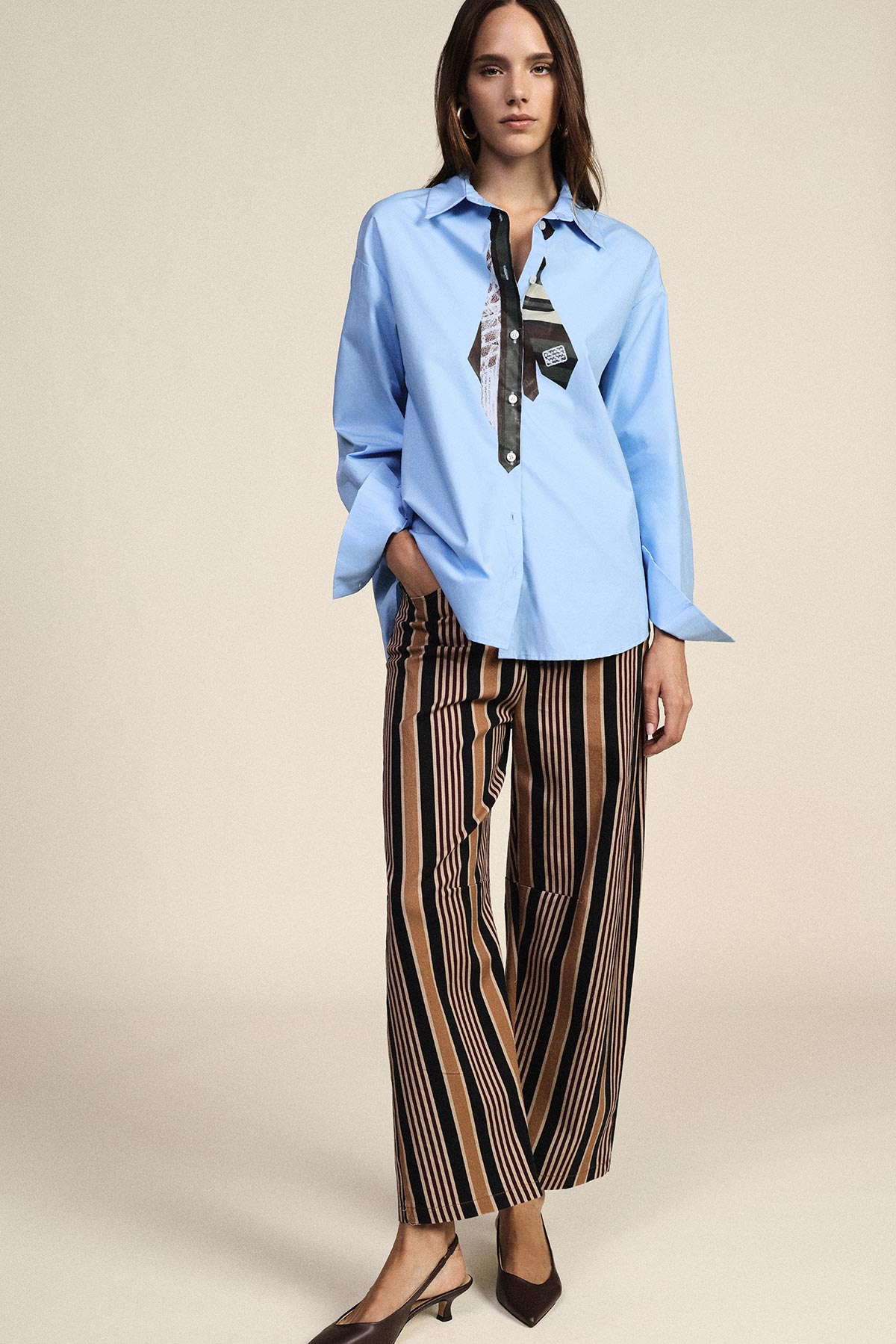 Woman modelling light blue shirt with patterned neck-tie, brown and black striped wide-leg trousers and low-heeled brown pumps on neutral background