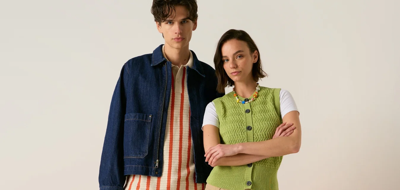 Giovane uomo e donna in piedi davanti a sedie arancioni, indossano abiti casual primaverili con sneakers, sfondo neutro.