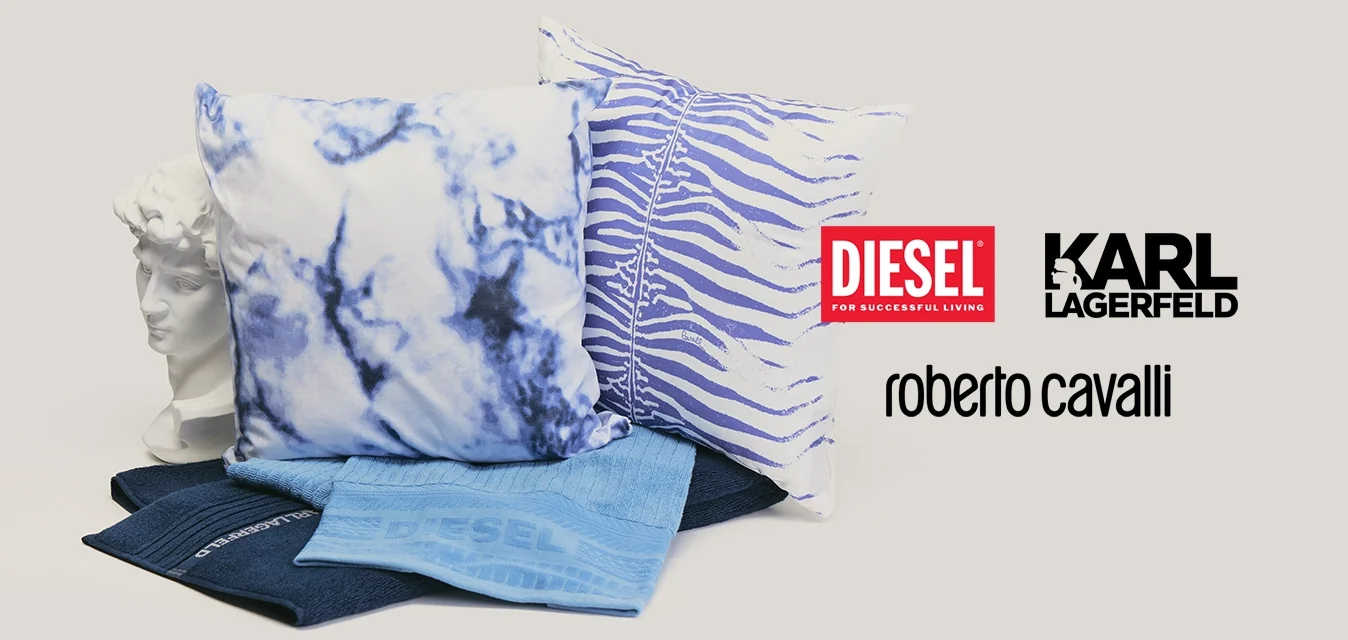 Due cuscini decorativi con motivi blu, due tessuti blu piegati e un busto bianco in stile classico su sfondo neutro.