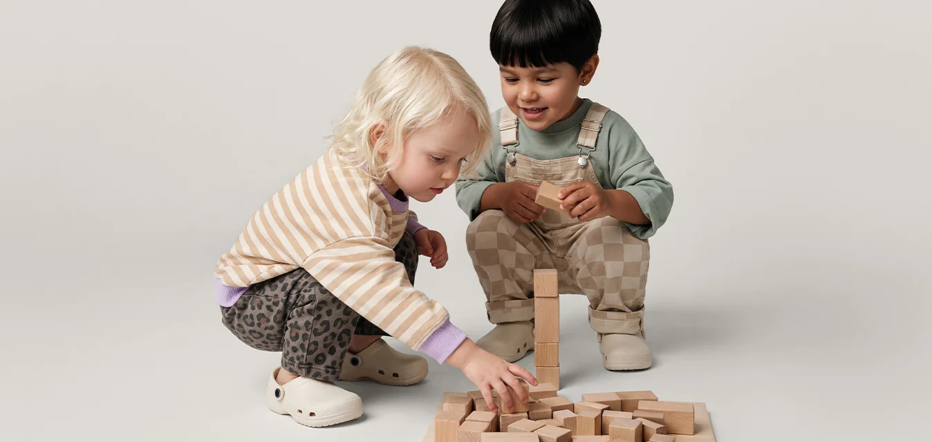 Due bambini piccoli giocano con blocchi di legno sul pavimento, costruendo insieme una piccola struttura su uno sfondo semplice.