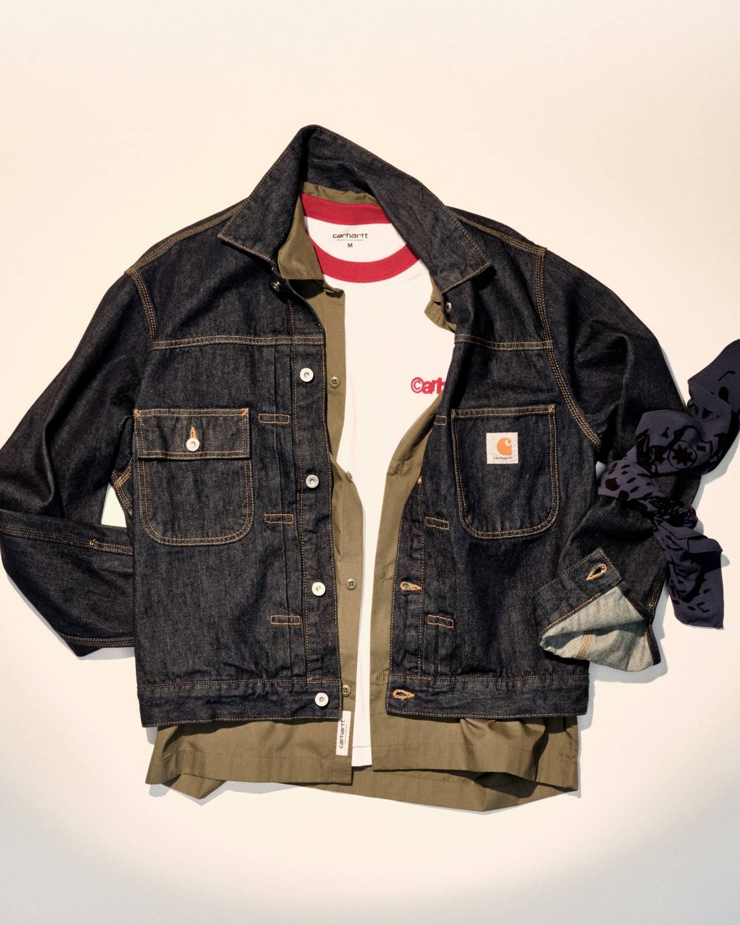 Dark denim Carhartt jacket layered over olive shirt and white tee with red ringer, styled flat with navy patterned scarf