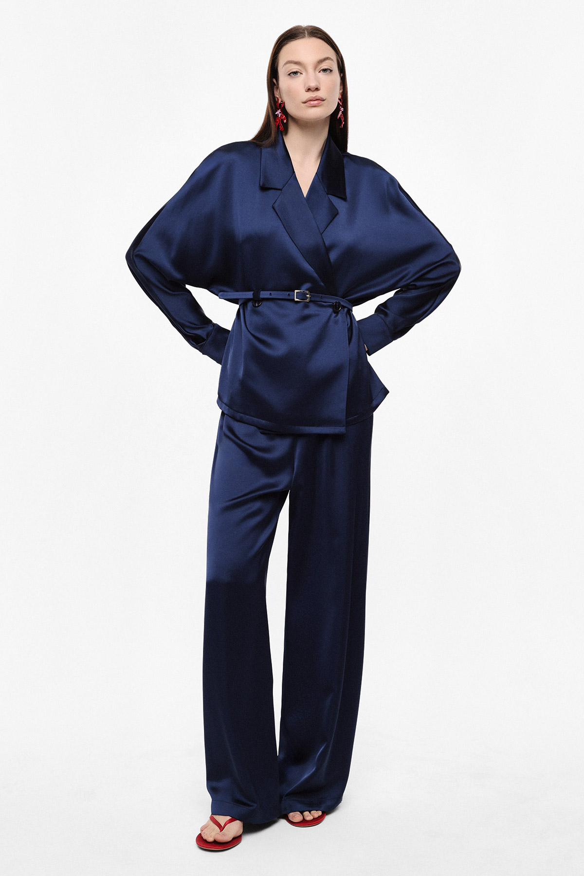 Model in navy satin belted blazer and wide-leg trousers with red earrings and sandals on white background