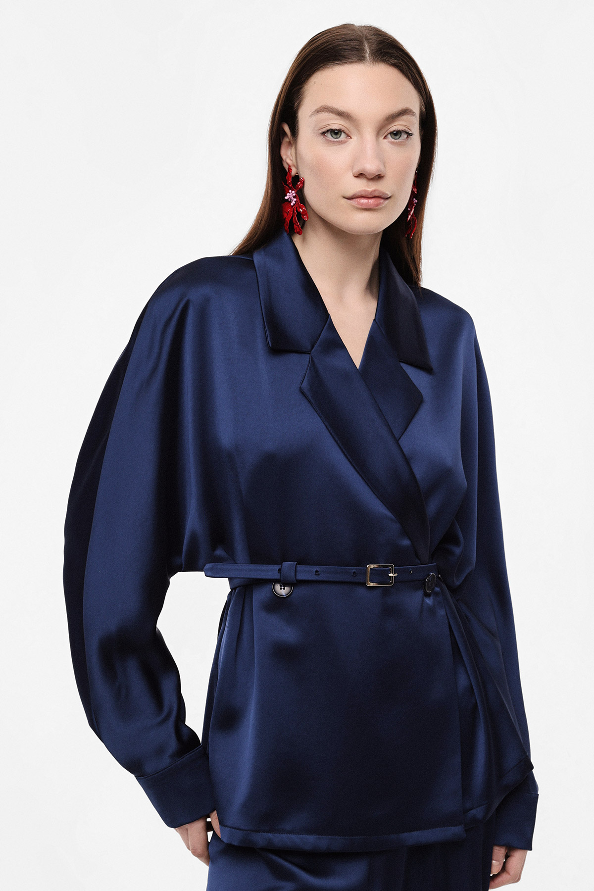Model wearing navy satin belted blazer with red statement earrings on white background