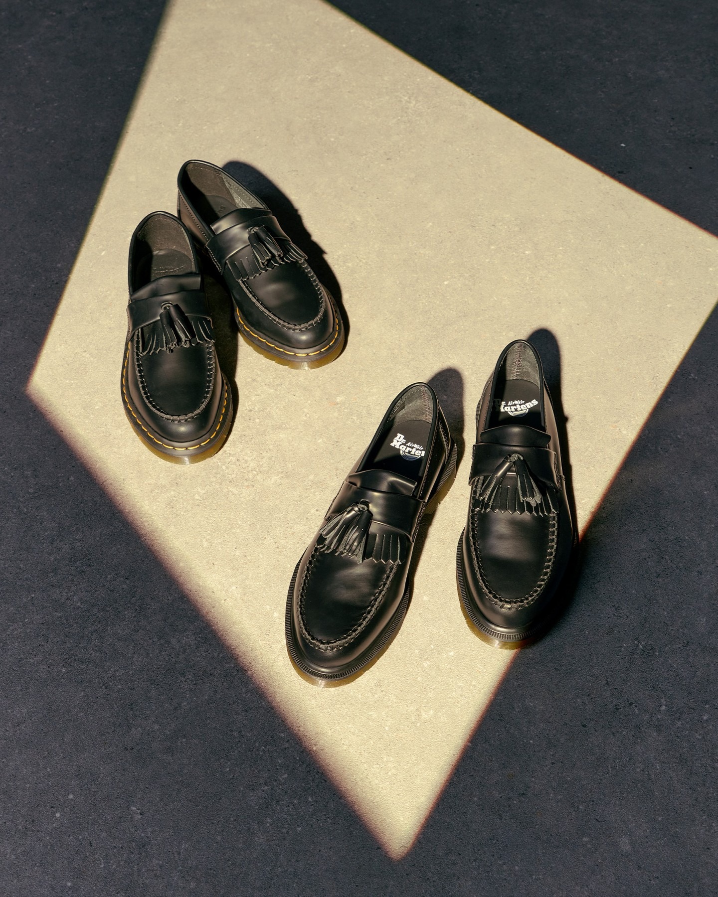 Two pairs of black leather tassel loafers displayed in sunlight on a beige square against dark flooring, top-down product flat lay