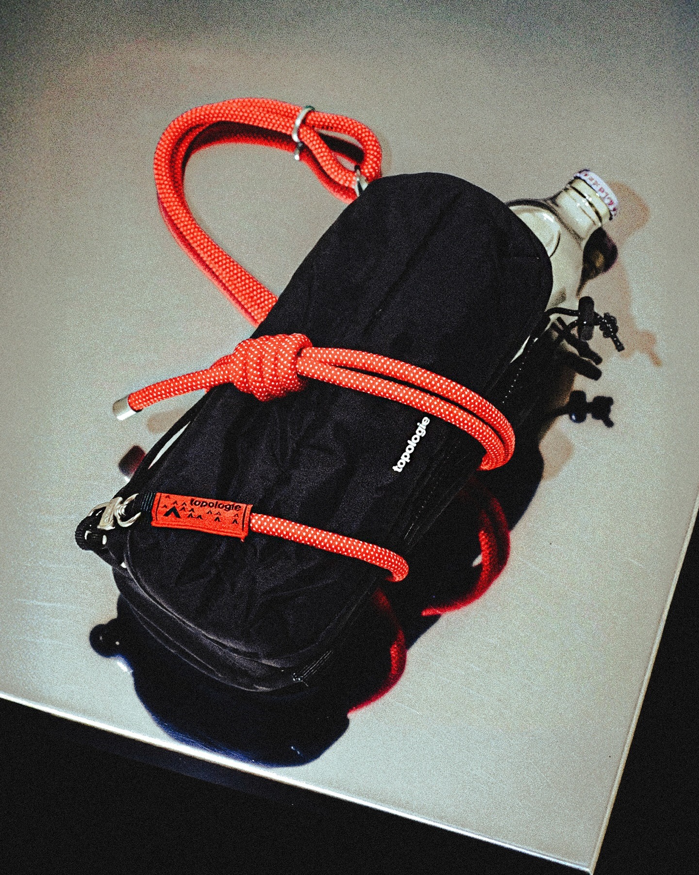 Black Topologie pouch secured with bright red climbing rope strap and a glass water bottle on a reflective tabletop