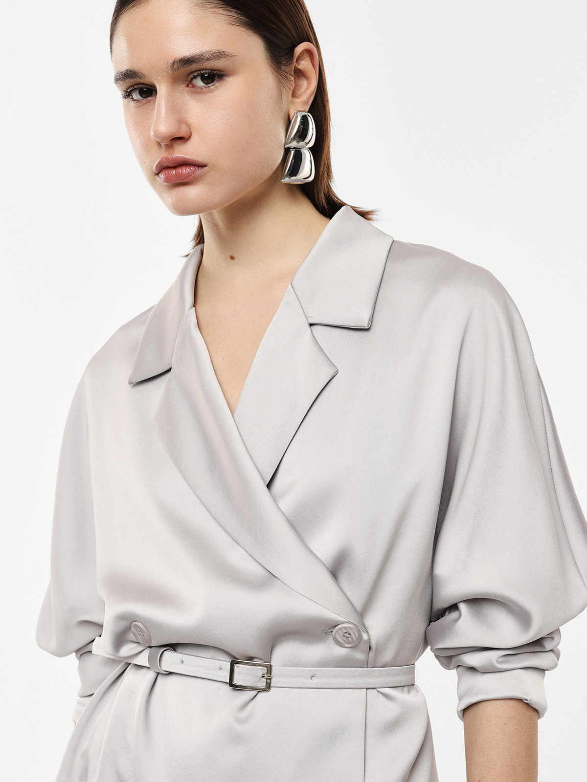 Young woman wearing pale grey belted wrap dress with rolled sleeves and large silver statement earrings against a neutral background