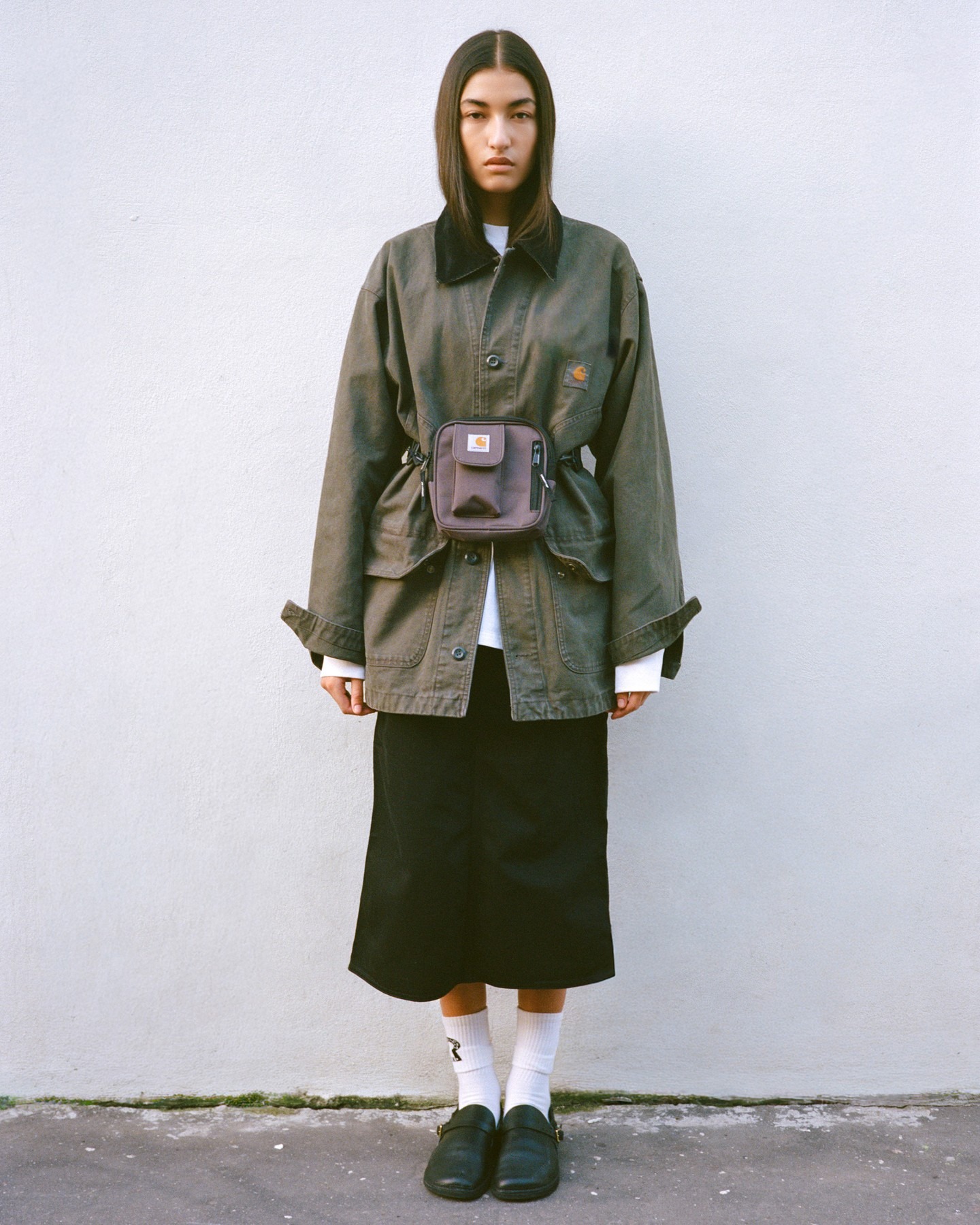 Young person standing against pale wall wearing oversized olive work jacket, small chest bag, black midi skirt, white socks and black shoes